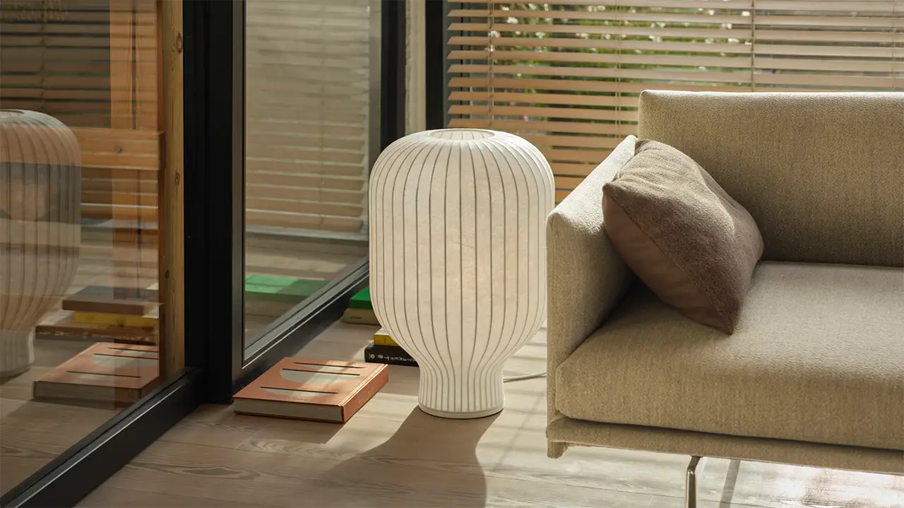 **Alt-Text:** Skandinavisch eingerichteter Raum mit einer weißen Muuto-Leuchte in organischer Form neben einem modernen Sofa, natürlichem Tageslicht und minimalistischer Holzoptik.