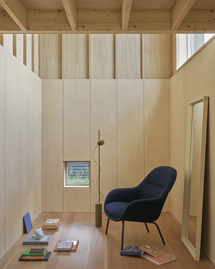 Muuto Post Leuchte **Alt-Text:**
Minimalistisch eingerichteter Raum mit hellem Holzinterieur, schwarzem Design-Sessel, Muuto Stehleuchte in Olivgrün und verstreut liegenden Büchern auf dem Boden – Licht fällt durch ein kleines Fenster und ein Oberlicht ein.