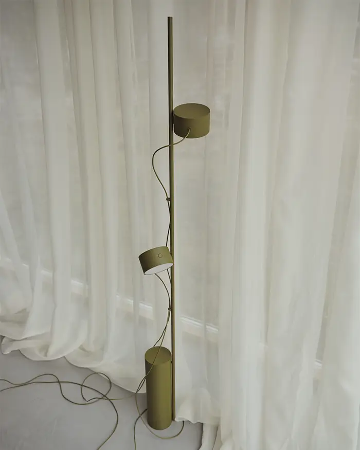 Muuto Post **Alt-Text:**
Moderne Stehleuchte von Muuto in Olivgrün mit drei verstellbaren Leuchtenköpfen an einer schlichten vertikalen Stange, platziert vor einem lichtdurchfluteten weißen Vorhang.