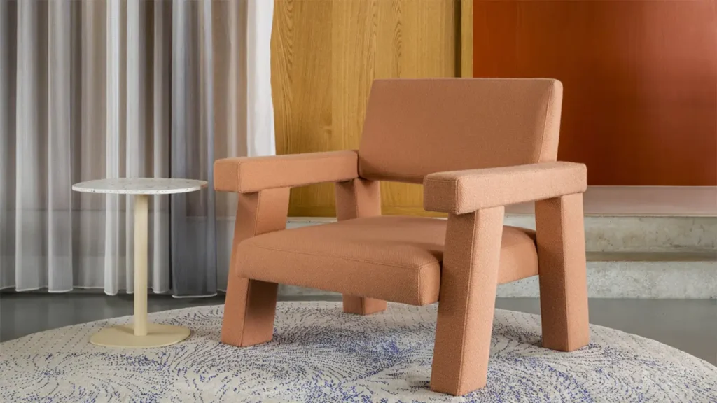 Der Mohs Chair von Philip Bogaerts in terracottafarbenem Wollbezug, zentral platziert vor Holzpaneelen und Vorhängen, neben einem runden Beistelltisch auf gemustertem Teppich – minimalistisches, skulpturales Design für zeitgemäße Räume.