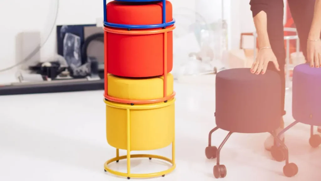 Drei farbenfrohe Drop Hocker von COR platzsparend gestapelt im Vordergrund, daneben zwei Rollhocker in Schwarz und Blau mit Händen in der Anwendungssituation – ideal für flexible Arbeitsbereiche.