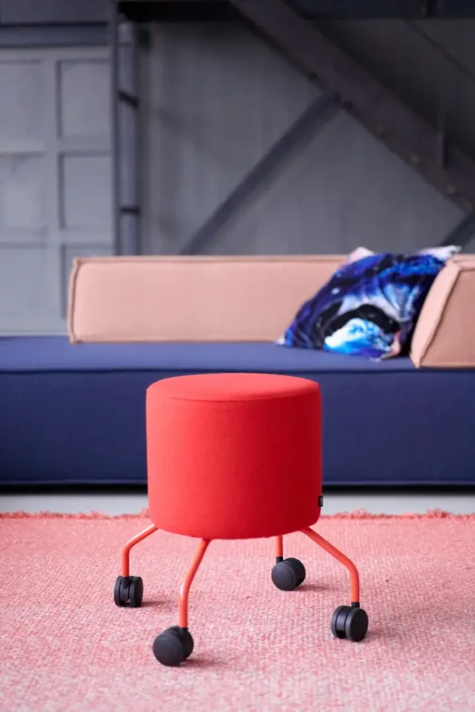 Ein roter Drop Rollhocker von COR auf rosa Teppich vor einer modernen Couch – kompakt, mobil und ideal als flexibler Sitzplatz im Wohn- oder Arbeitsbereich.