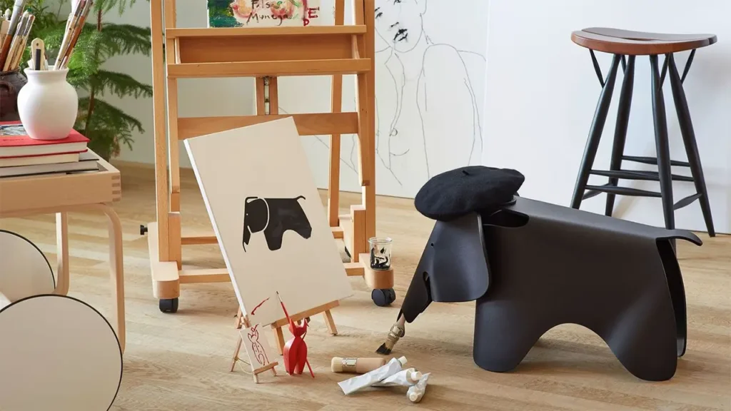Schwarzer Eames Elephant von Vitra mit Baskenmütze in einem Atelier-Setting, vor einer Staffelei mit Elefantenbild und Malutensilien – humorvolle Inszenierung des Designklassikers.