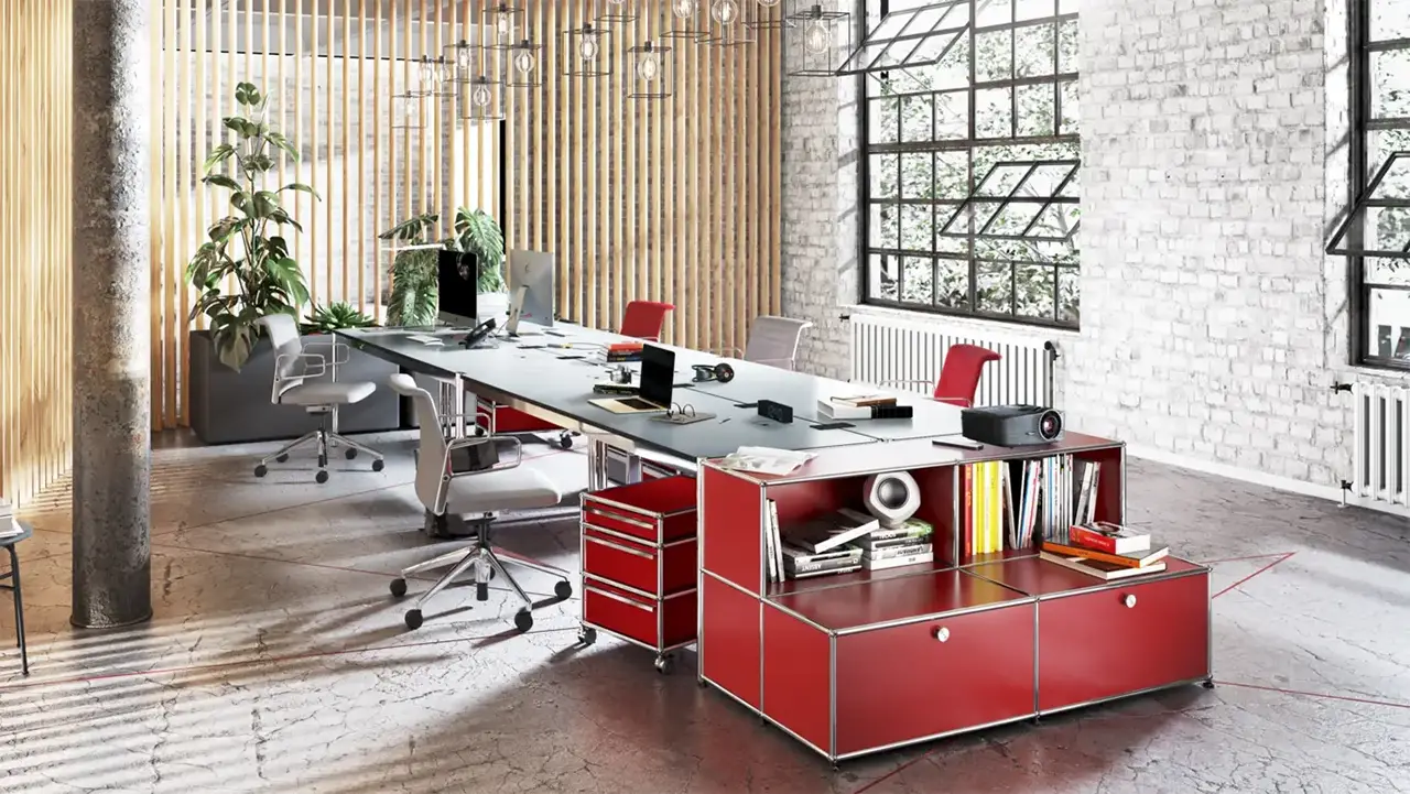 Helles, modernes Büro mit industriellem Charme, ausgestattet mit einem langen Arbeitstisch, roten USM Haller Rollcontainern und Stauraummodulen. Große Fenster, rote Akzente, Designstühle und grüne Pflanzen schaffen eine inspirierende Arbeitsatmosphäre.