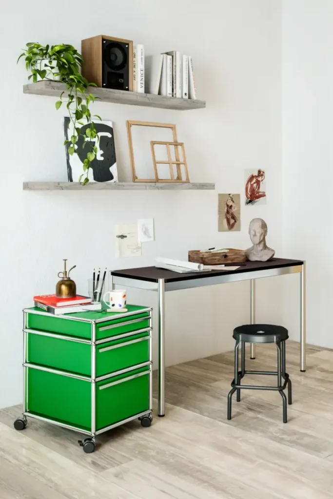 Künstlerisches Arbeitszimmer mit grünem USM Haller Rollcontainer Kreativ eingerichtetes Homeoffice mit minimalistischem Tisch, grünem USM Haller Rollcontainer und schwarzen Hocker. An der Wand hängen Zeichnungen und Skizzen, darüber zwei rustikale Holzregale mit Büchern, Pflanzen und Dekoobjekten. Ein inspirierender Ort für Kunst und Ideen.
