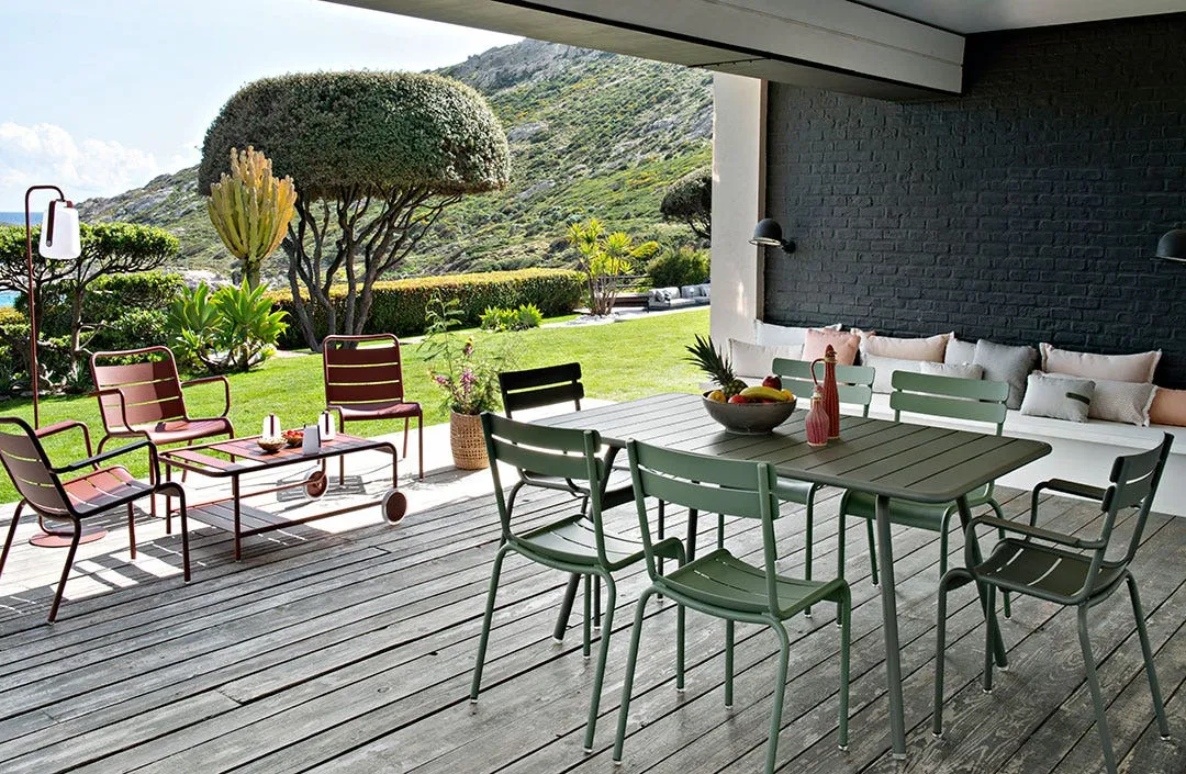 Überdachte Terrasse mit grünem Esstisch-Set und roten Lounge-Stühlen der Fermob Luxembourg-Kollektion, im Hintergrund mediterraner Garten mit Bergkulisse
