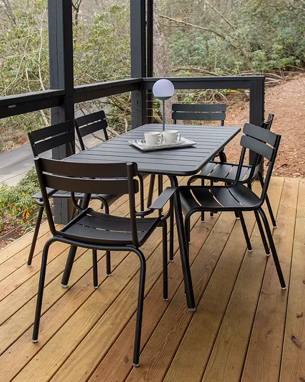 Schlichte Eleganz mit Fermob Luxembourg in Schwarz auf Holzterrasse Schwarzes Essgruppen-Set der Fermob Luxembourg-Kollektion auf überdachter Holzterrasse im Wald, mit sechs Stühlen und rechteckigem Tisch, dekoriert mit Tablett und Outdoor-Leuchte