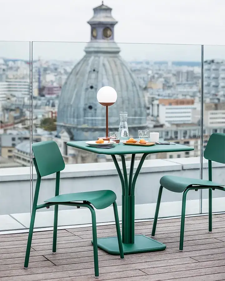 Urbaner Ausblick mit Fermob: Rooftop-Setting über den Dächern von Paris Grünes Bistrotisch-Set von Fermob auf einer Dachterrasse mit Glasgeländer, Blick auf Pariser Kuppelbau, Frühstücksgedeck und dimmbare Outdoor-Leuchte bei bedecktem Himmel
