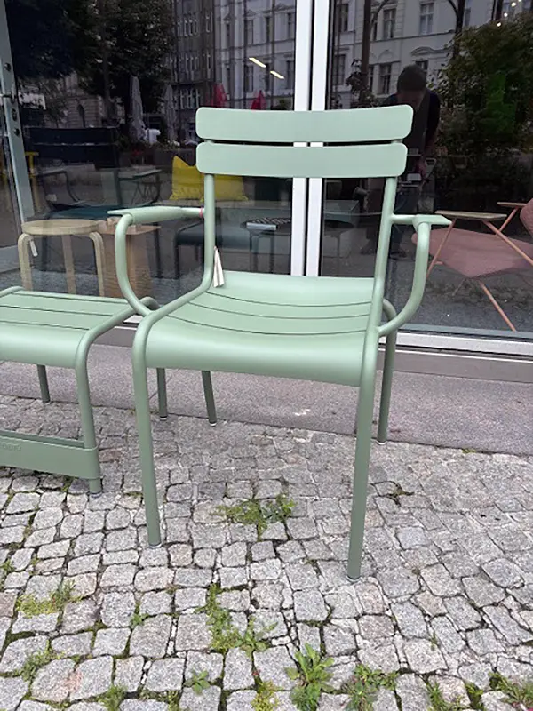 Fermob Luxembourg Armlehnstuhl in Salbeigrün vor dem Showroom Grüner Luxembourg-Armlehnstuhl von Fermob auf Pflastersteinen vor einer Glasfassade mit Blick in den Showroom von UnitedSpaces in Berlin