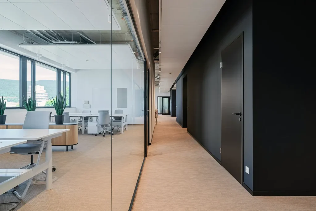 Moderner Büroflur mit schwarzer Wandgestaltung, Glasfront und Blick in einen hellen Arbeitsbereich mit weißen Schreibtischen, grauen Bürostühlen und großen Fenstern.