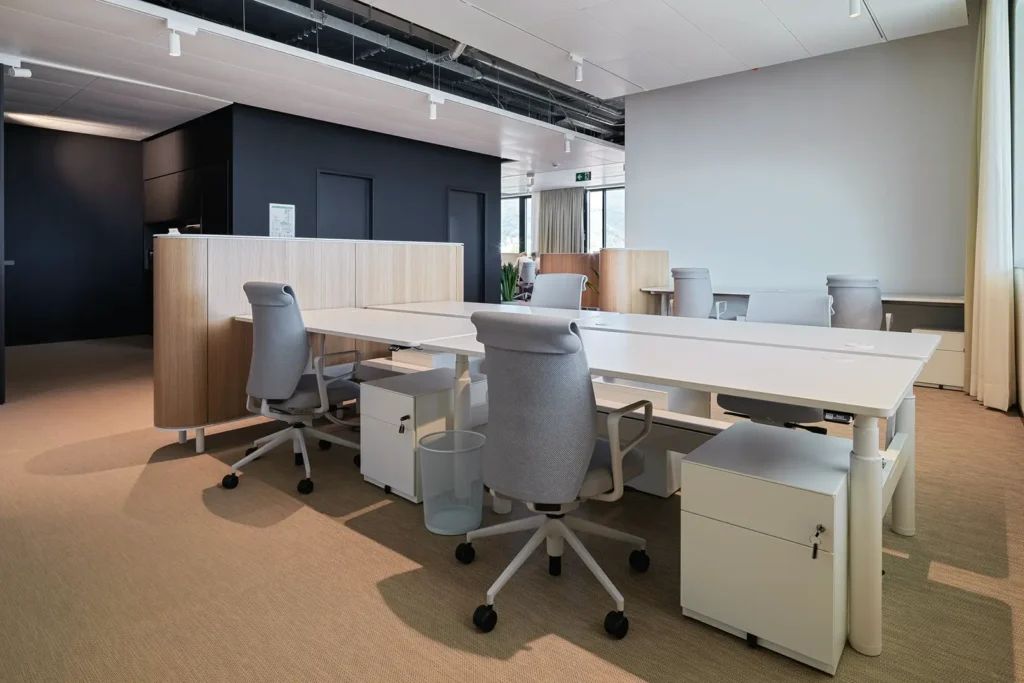 Moderner Arbeitsbereich mit weißen Schreibtischen, grauen ergonomischen Bürostühlen, rollbaren Container-Schränken und Holztrennelementen vor schwarzer Wandgestaltung.