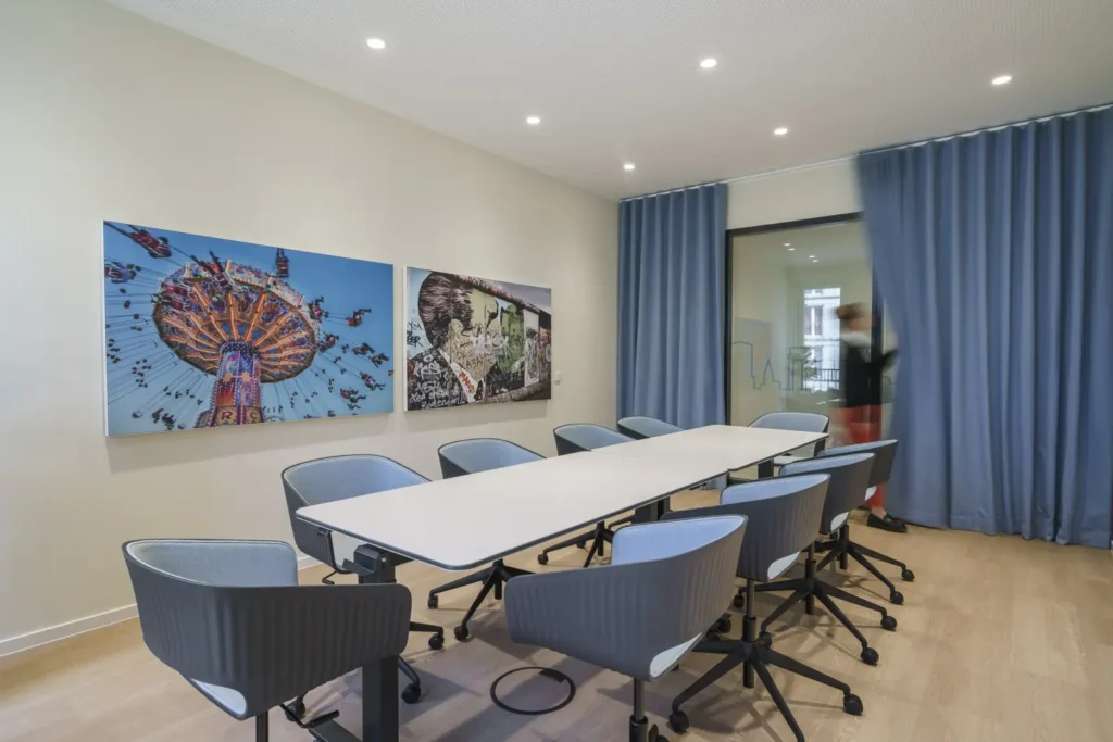 Heller Besprechungsraum mit langem weißen Tisch, grauen Drehstühlen, blauen Vorhängen und zwei farbenfrohen Kunstwerken an der Wand.