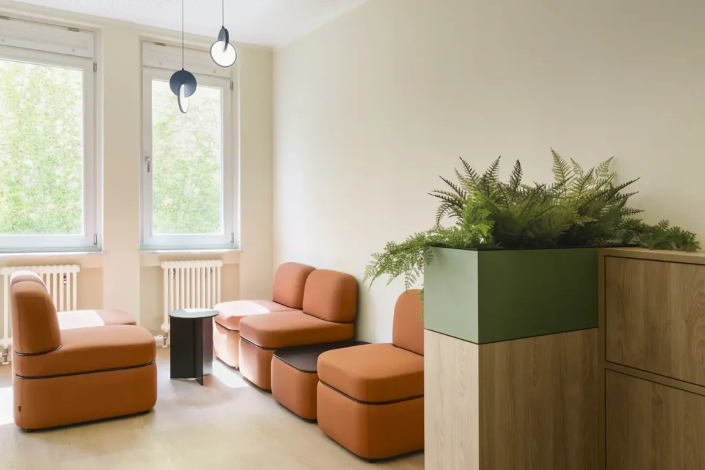 Heller Raum mit bodentiefen Fenstern, modernen orangefarbenen Lounge-Sesseln, kleinem Beistelltisch und einem grünen Pflanzgefäß auf Holzpodest.