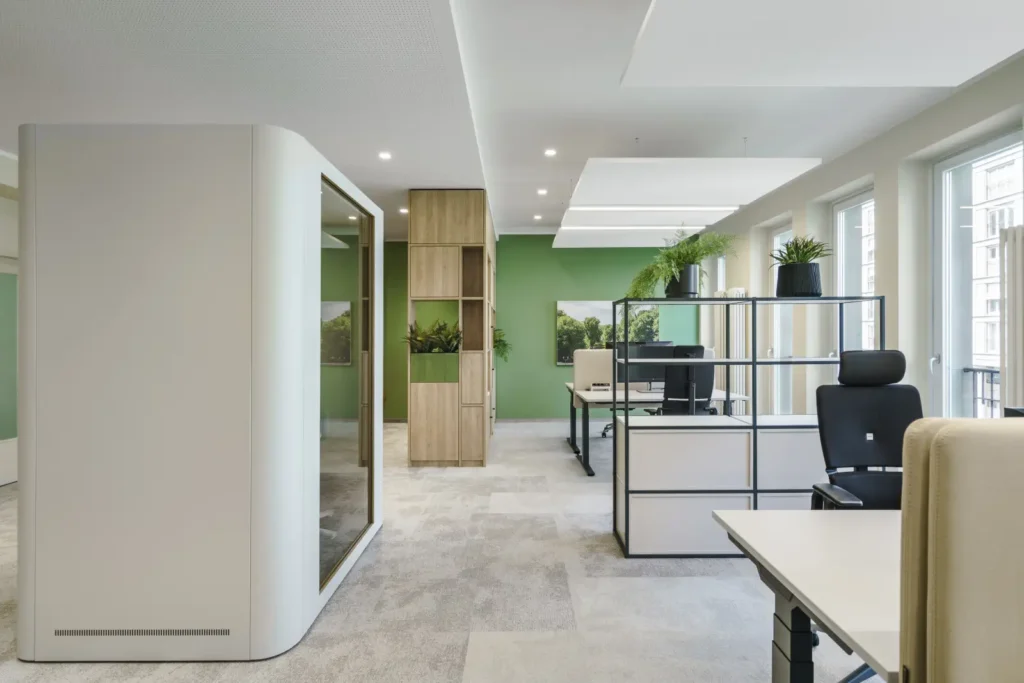 Offener Bürobereich mit großen Fenstern, grünen Wänden, modernen Arbeitsplätzen, Regal mit Pflanzen und schallisolierter Meetingbox.