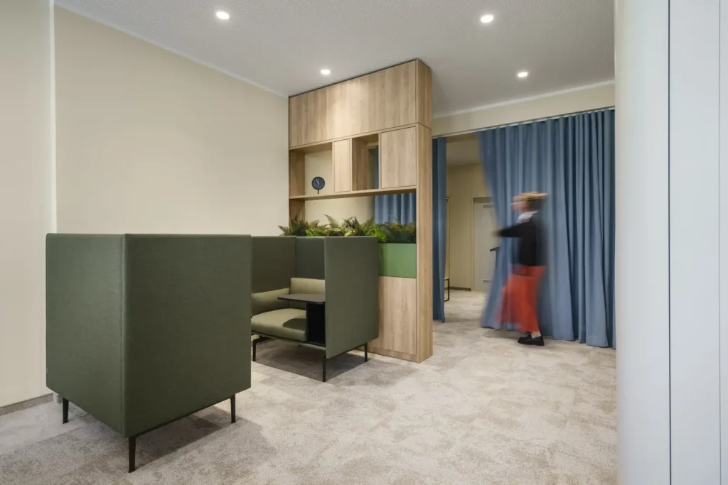 Moderner Arbeitsbereich mit hohen grünen Akustiksesseln, integriertem Holzelement mit Pflanzen und Stauraum sowie blauem Vorhang im Hintergrund.
ChatGPT fragen