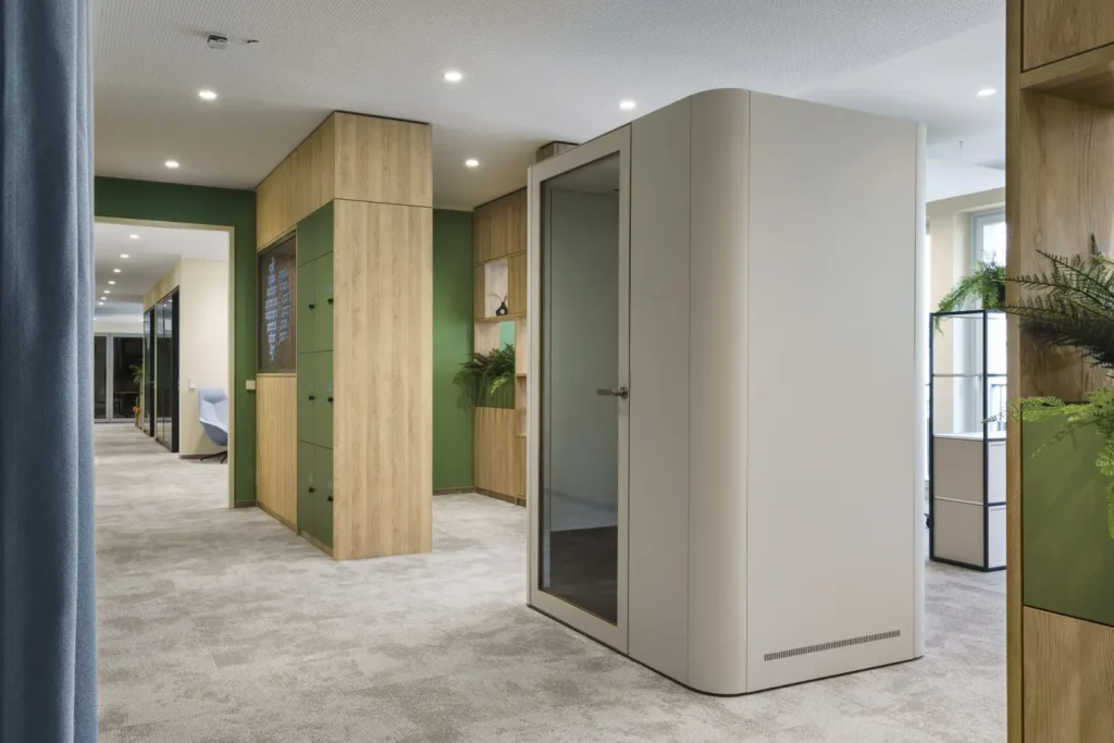 Heller Büroflur mit freistehender Telefon- bzw. Meetingbox, grünen und holzverkleideten Schränken, dekorativen Pflanzen und Sicht auf weitere Arbeitsbereiche.
ChatGPT fragen